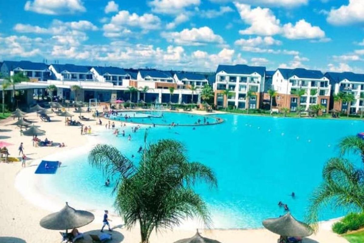 A large, resort-style swimming pool with a sandy beach area, surrounded by modern apartment buildings in a well-maintained residential complex.