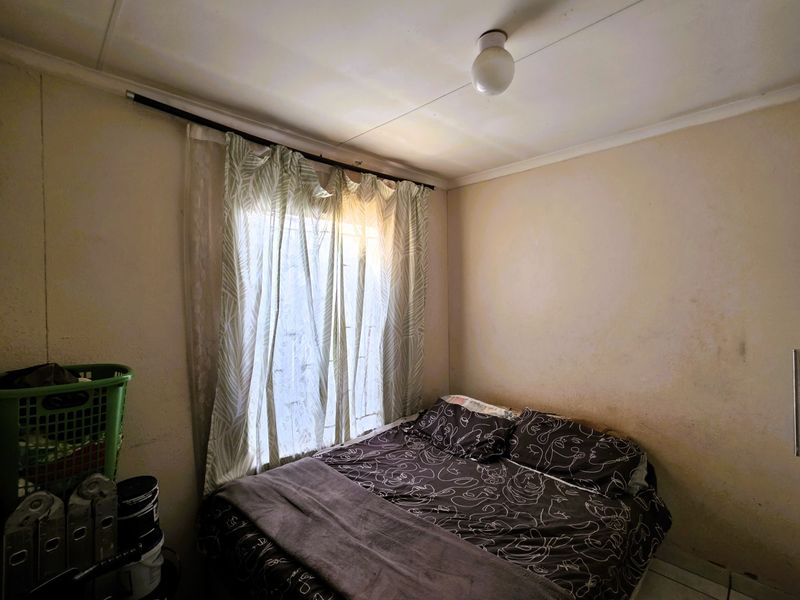 A compact bedroom featuring a window with curtain rod and basic ceiling lighting, requiring cosmetic updates to the wall finishes.
