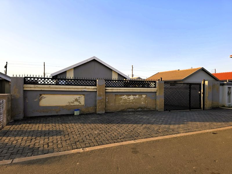 Front exterior view of the property featuring a paved driveway, metal security gate, and a boundary wall requiring maintenance and repainting.