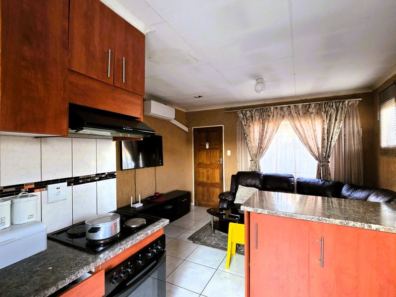 Open-plan kitchen and living area featuring wooden cabinetry, granite countertops, and tiled flooring, with visible ceiling and wall maintenance required.