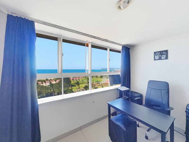 A bright office space featuring large sliding windows with expansive coastal and park views, tiled flooring, and neutral wall finishes.