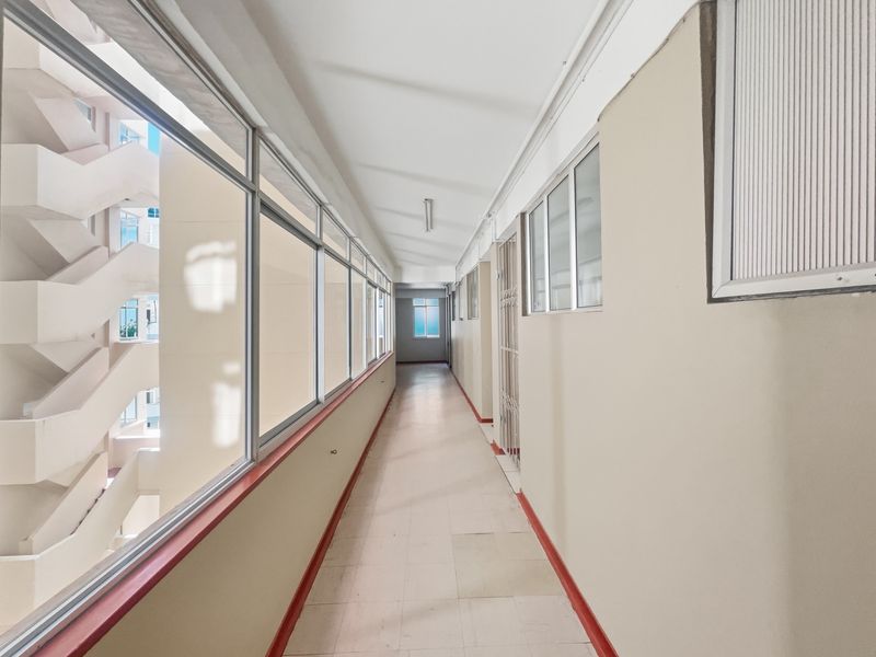 A long, well-lit apartment building hallway featuring large glass windows, tiled flooring, and a security gate, providing access to residential units.