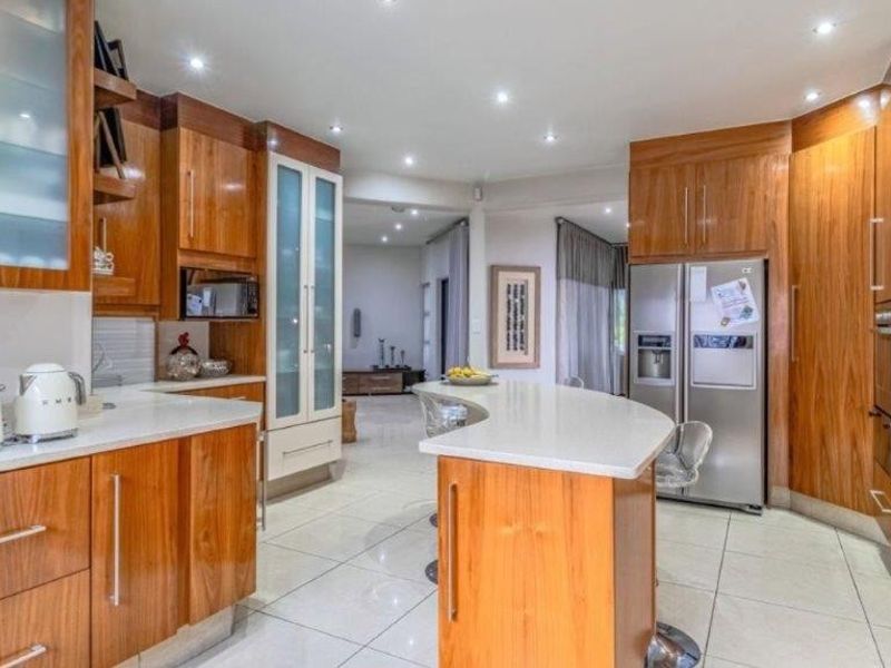 A well-maintained kitchen featuring warm-toned wooden cabinetry, stone countertops, a central island, and modern recessed lighting, flowing into an open-plan living area.