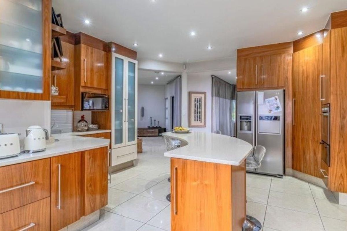 A well-maintained kitchen featuring warm-toned wooden cabinetry, stone countertops, a central island, and modern recessed lighting, flowing into an open-plan living area.