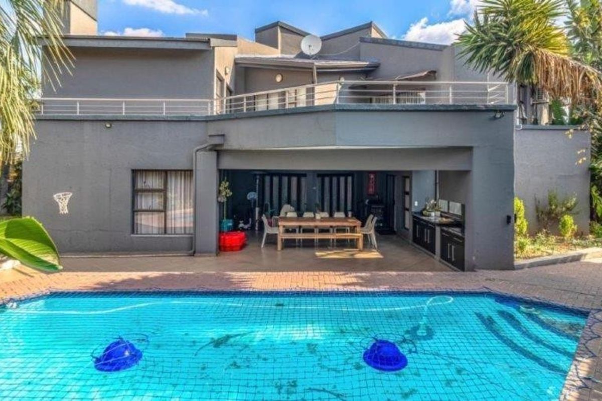 Rear exterior view featuring a swimming pool with safety net, a covered patio area with built-in braai, and an upper-level balcony.