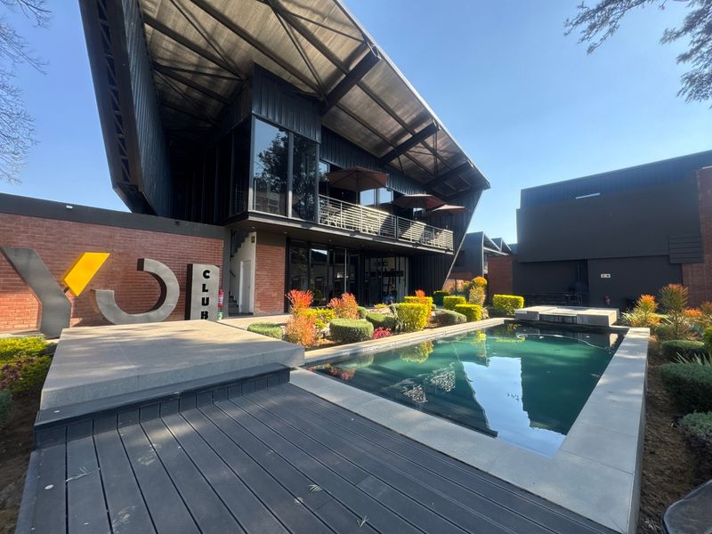 Modern clubhouse facility featuring a swimming pool, timber decking, landscaped gardens, and a striking architectural design with a prominent metal roof structure.