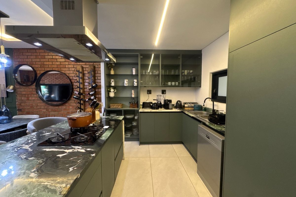 Modern kitchen featuring a stone island with gas hob, sleek green cabinetry, integrated shelving, and an exposed brick feature wall.