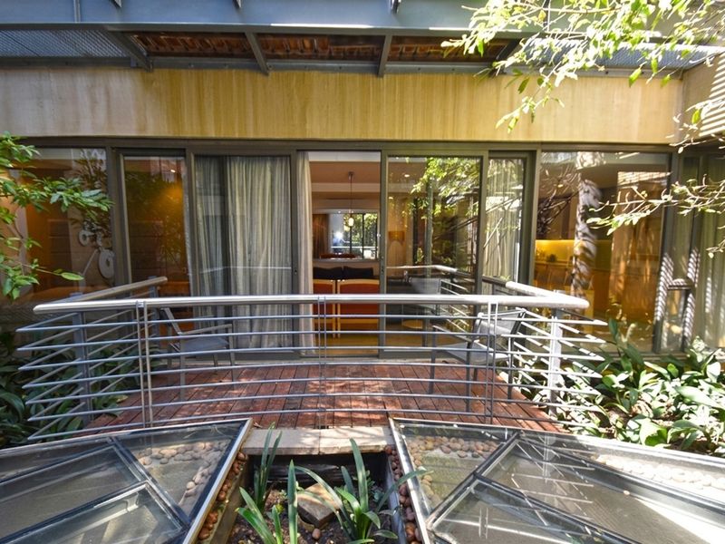A modern balcony featuring timber decking, a stainless steel balustrade, and a metal pergola, providing seamless indoor-outdoor flow through large sliding glass doors.