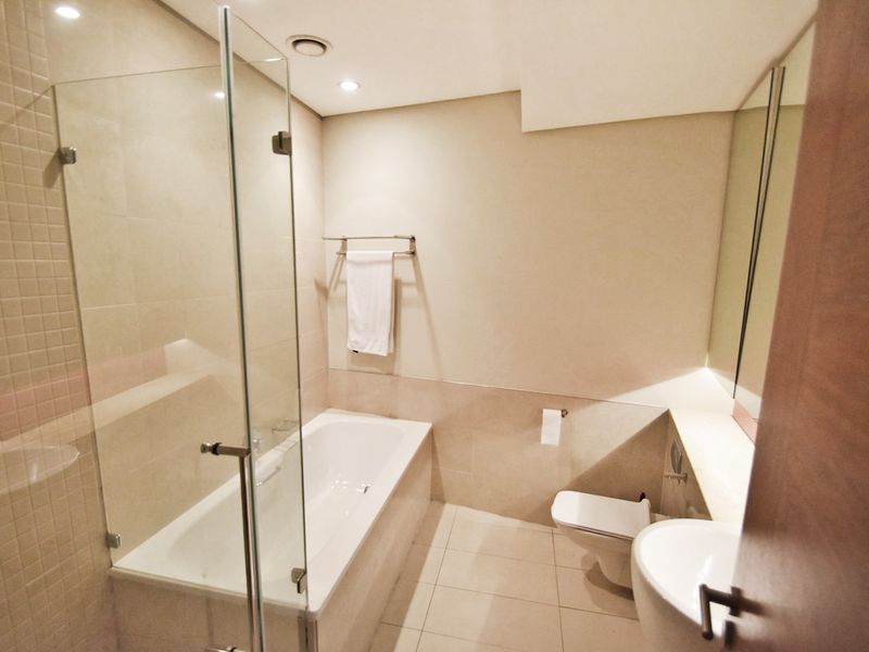 A modern, neutral-toned bathroom featuring a built-in bathtub with a glass screen, wall-mounted toilet, and a sleek vanity basin.