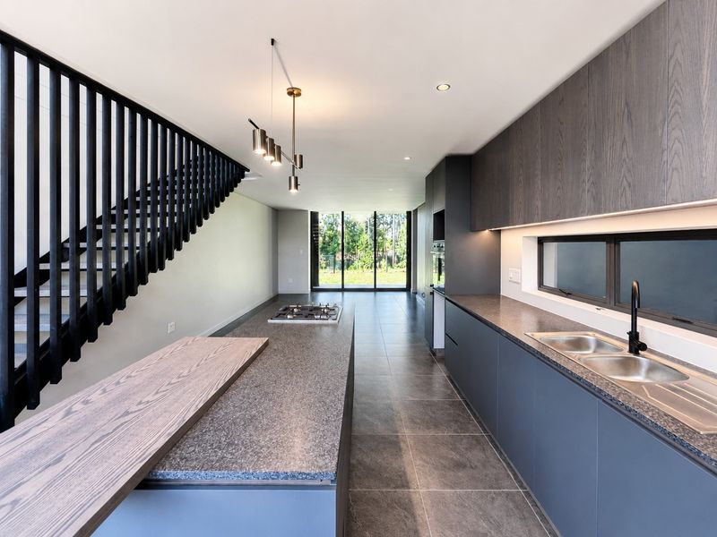 Modern open-plan kitchen featuring a large island with gas cooktop, sleek cabinetry, integrated appliances, and a striking staircase, leading to a bright rear garden view.