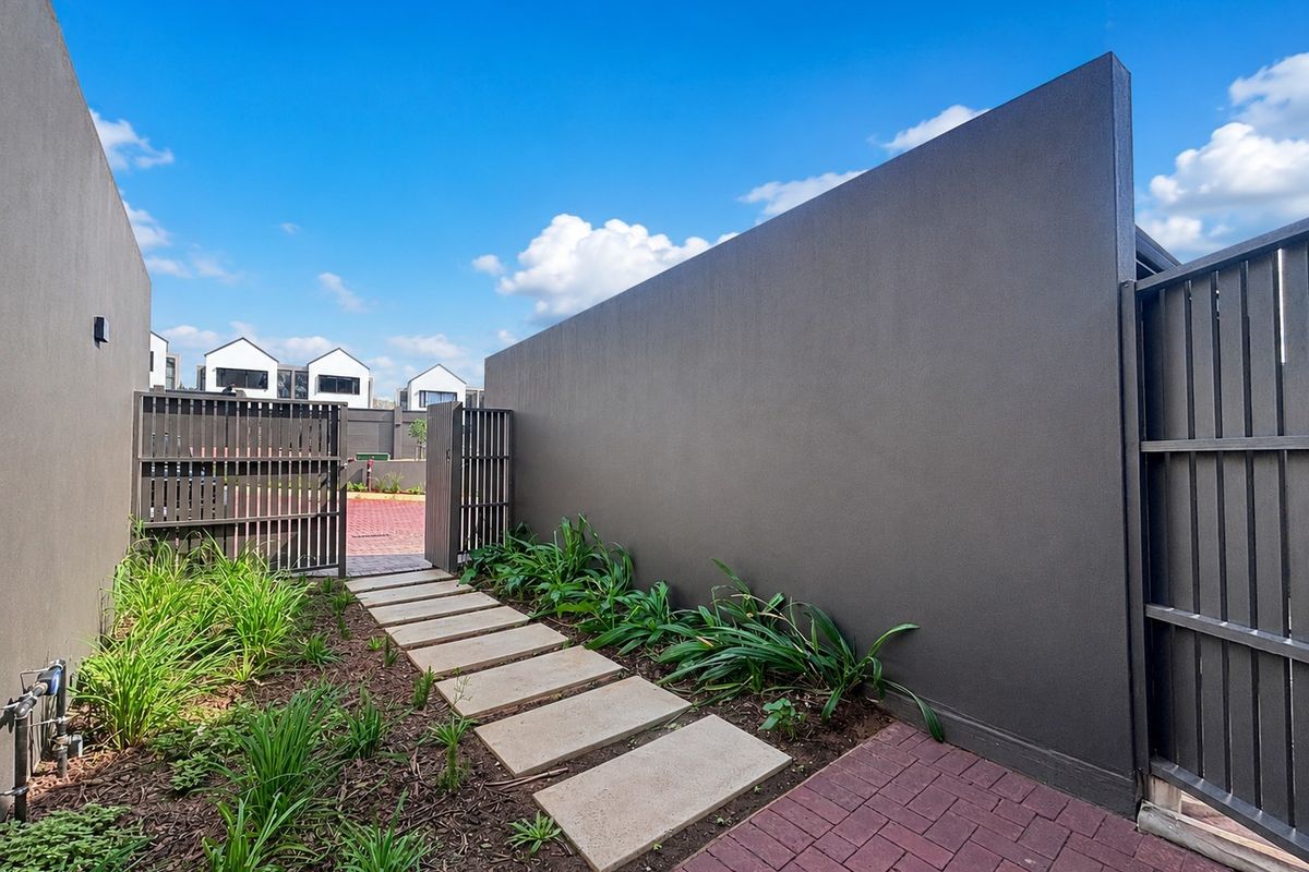 A well-maintained rear courtyard featuring a modern boundary wall, metal gate, brick paving, and a stone pathway leading through landscaped garden beds.