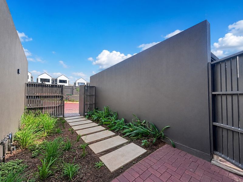 A well-maintained rear courtyard featuring a modern boundary wall, metal gate, brick paving, and a stone pathway leading through landscaped garden beds.