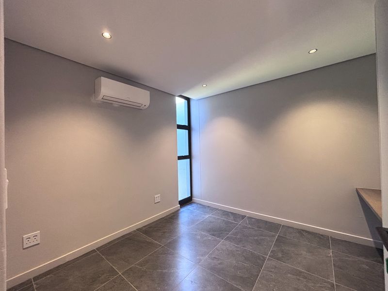 A modern, empty bedroom featuring tiled flooring, recessed downlighting, a built-in desk, and a wall-mounted air conditioning unit.