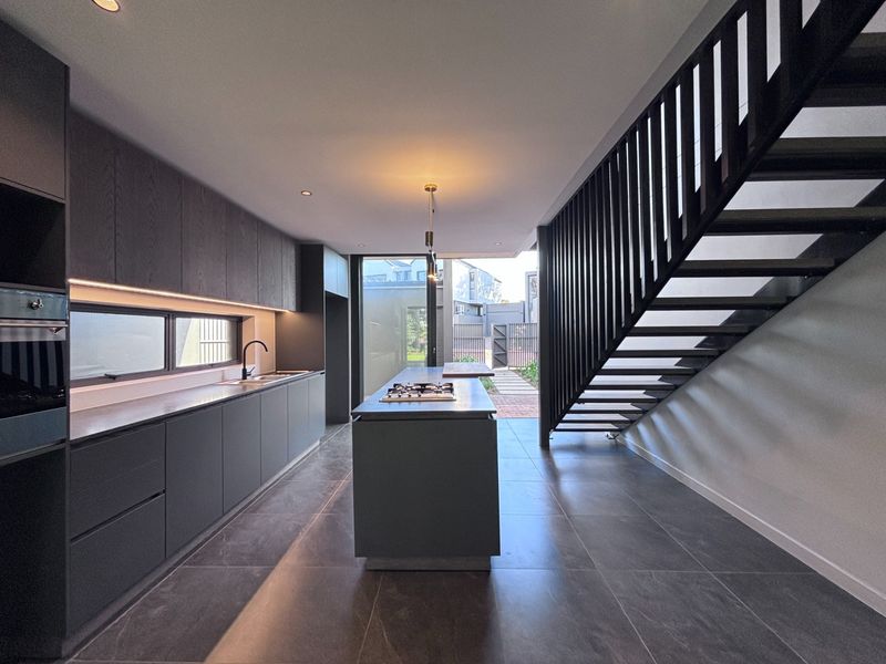 Modern open-plan kitchen featuring dark cabinetry, a central island with a gas cooktop, and sleek tiled flooring, adjacent to a contemporary staircase.