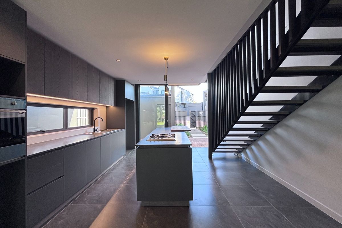 Modern open-plan kitchen featuring dark cabinetry, a central island with a gas cooktop, and sleek tiled flooring, adjacent to a contemporary staircase.
