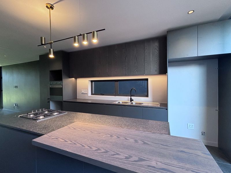 Modern kitchen featuring dark wood cabinetry, stone countertops, integrated gas cooktop, built-in oven, and contemporary pendant lighting in an open-plan layout.