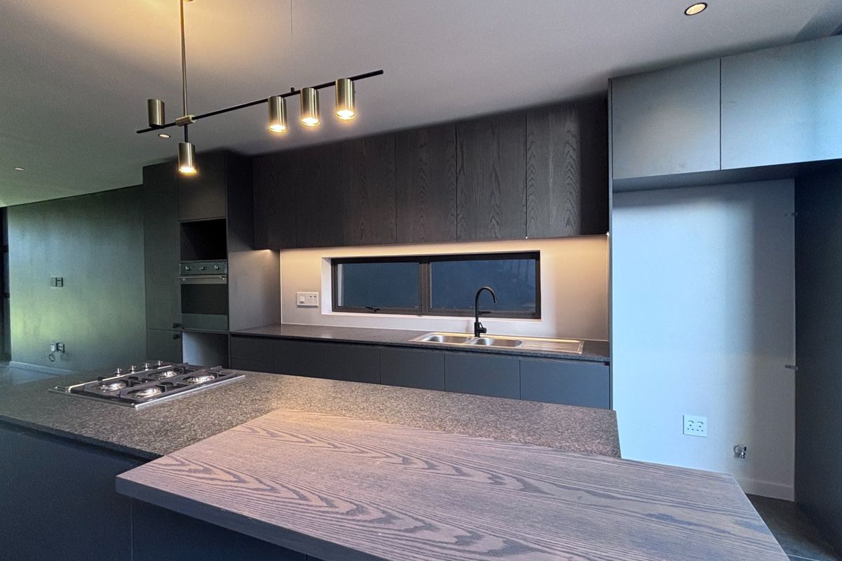 Modern kitchen featuring dark wood cabinetry, stone countertops, integrated gas cooktop, built-in oven, and contemporary pendant lighting in an open-plan layout.