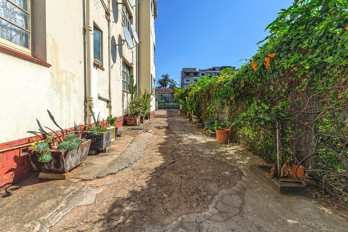 A narrow exterior rear walkway featuring cracked concrete paving, perimeter fencing with climbing vegetation, and exterior building wall with drainage infrastructure.
