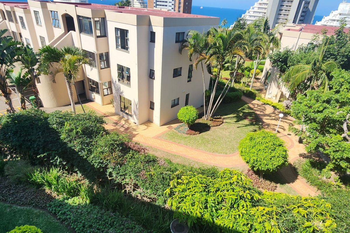 An elevated aerial view of the apartment complex grounds, featuring well-maintained landscaped gardens, paved pathways, and surrounding residential buildings.