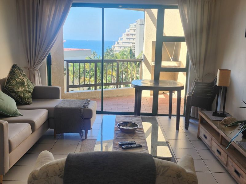 A comfortable living room featuring tiled flooring and a large sliding glass door that opens onto a balcony with scenic coastal views.
