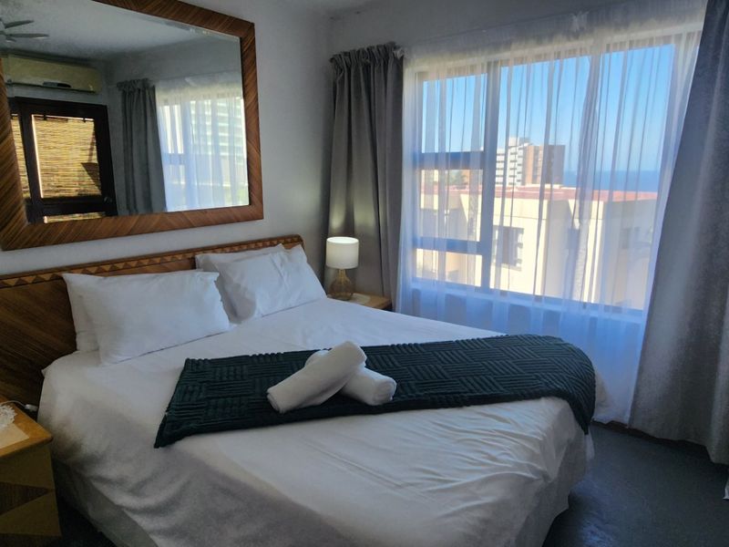 A comfortable bedroom featuring a large window with natural light, a wall-mounted mirror, and an integrated air conditioning unit.