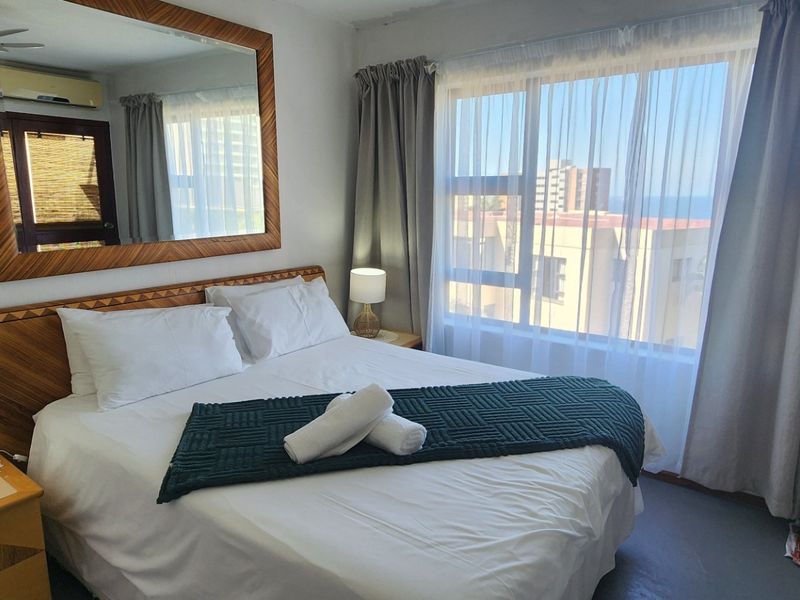 A well-lit bedroom featuring a large window with natural light, a wall-mounted mirror, and an integrated air conditioning unit.