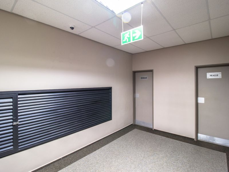 A clean commercial hallway featuring suspended ceilings, security surveillance, emergency signage, and access doors to male and female restroom facilities.