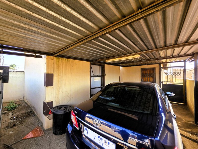 A covered carport area featuring a corrugated metal roof, security gate, and steel window frames, providing sheltered parking space.