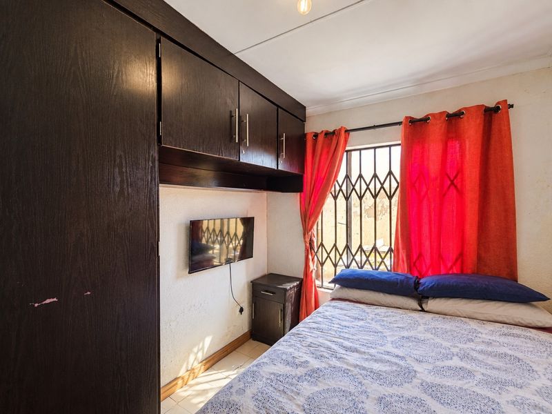 A compact bedroom featuring built-in dark wood cabinetry, tiled flooring, and a window fitted with a security gate.