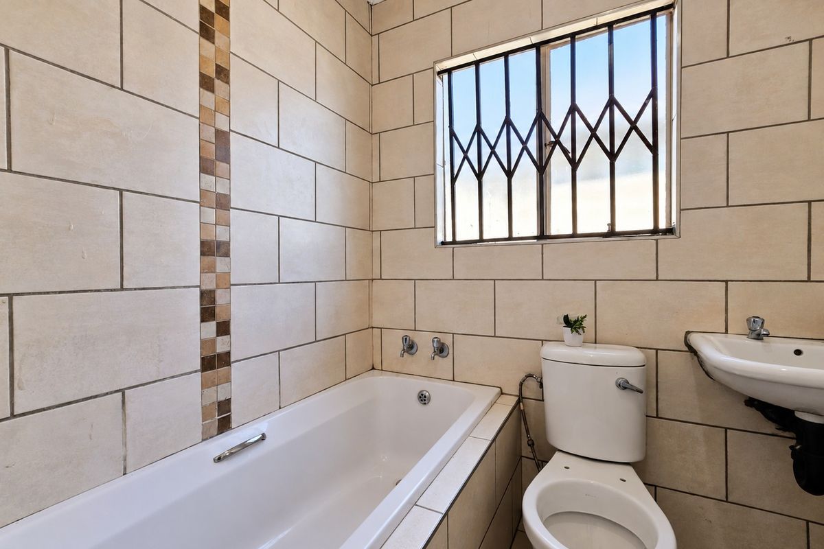 A functional bathroom featuring a built-in bathtub, wall-mounted basin, and toilet, finished with neutral ceramic wall tiles and security bars on the window.