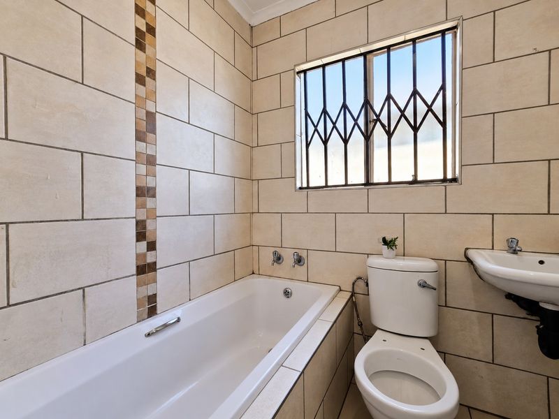 A functional bathroom featuring a built-in bathtub, wall-mounted basin, and toilet, finished with neutral ceramic wall tiles and security bars on the window.