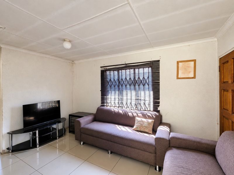 A living room featuring tiled flooring, a security gate on the window, and a timber door, showing signs of wear on the ceiling and walls.