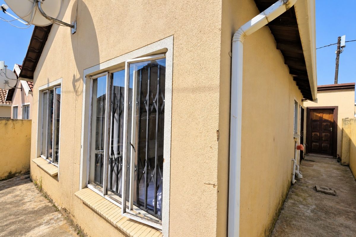 Rear exterior view of the property featuring plastered walls, security gates on windows, and concrete paving with visible signs of weathering and maintenance needs.