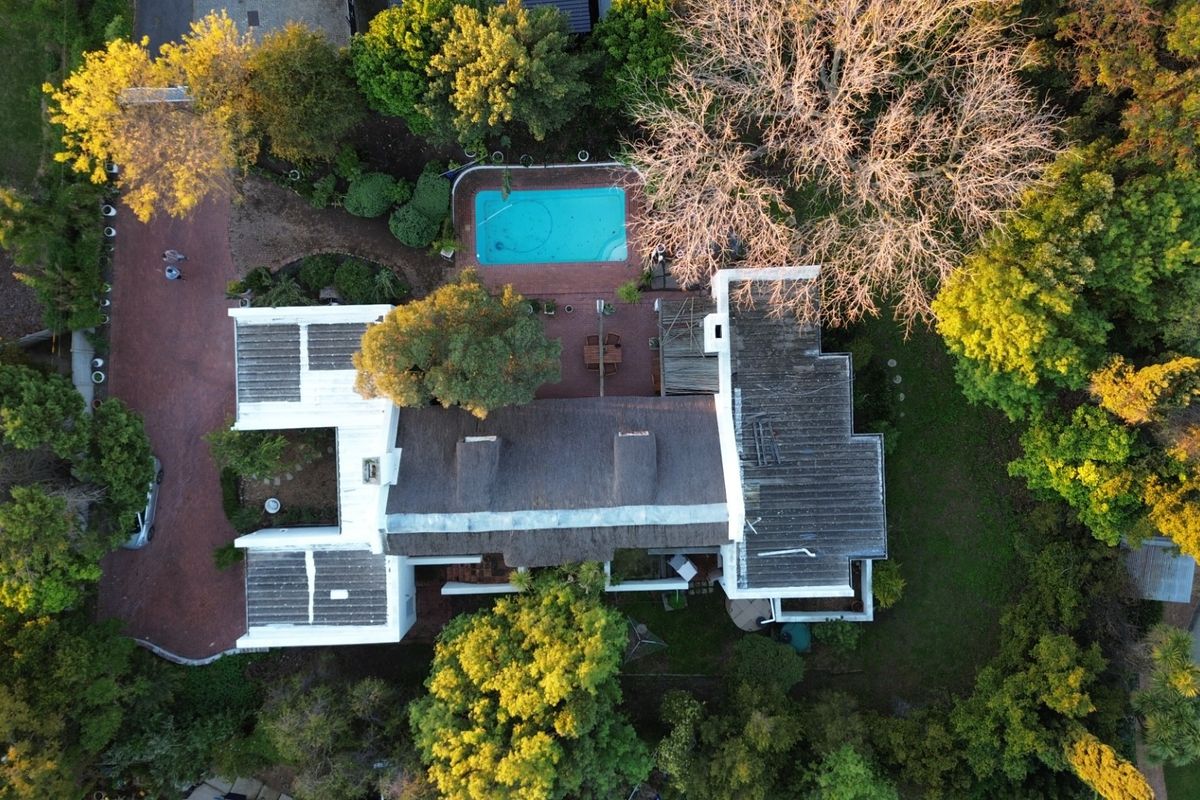 Aerial view of the property showcasing the main residence with a thatched roof, a swimming pool, paved driveway, and established garden surroundings.