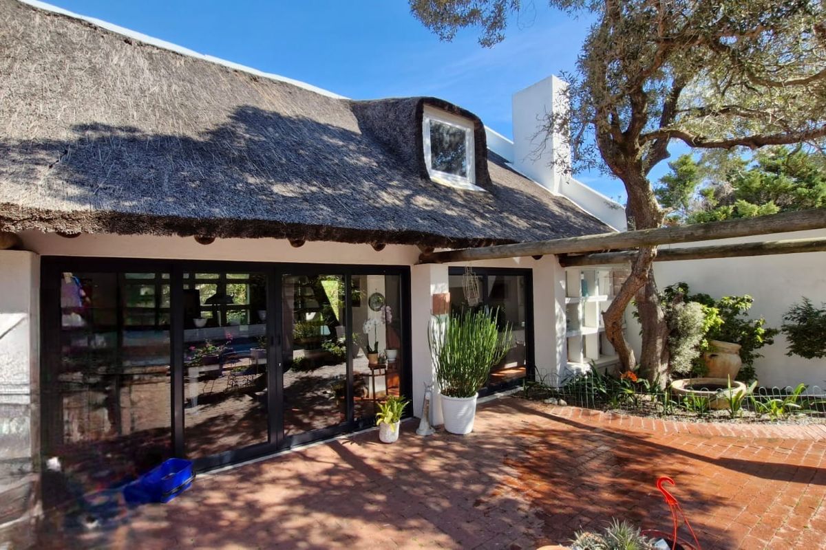 Rear exterior view of a charming thatched-roof house featuring a brick patio, timber pergola, and large sliding glass doors leading to the interior.