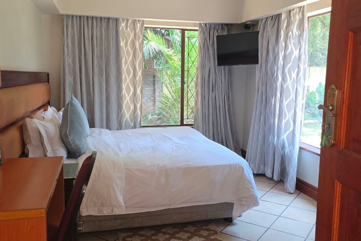 A well-maintained guest bedroom featuring tiled flooring, a built-in headboard, and large windows with curtains overlooking the garden.