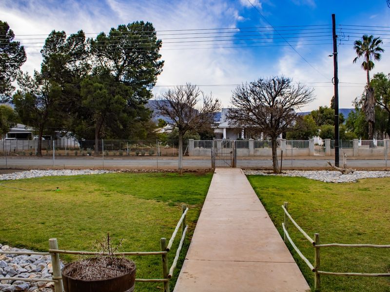 A view of the front garden featuring a concrete pathway leading to a gated entrance with established trees and perimeter fencing.