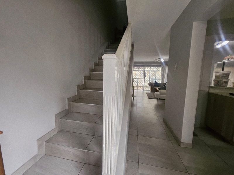 A tiled hallway featuring a staircase with a white wooden balustrade, leading towards an open-plan living area with natural light.