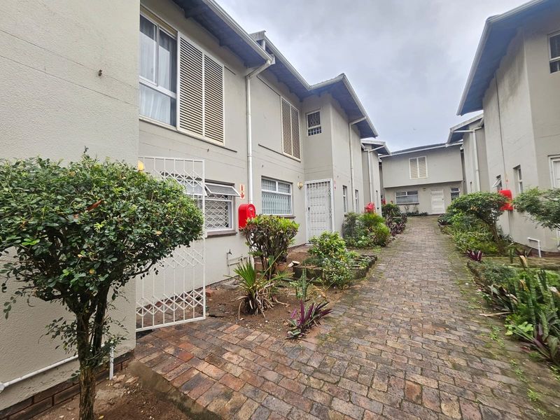 A view of the rear exterior walkway of the townhouse complex, featuring brick paving, security gates, and established garden beds.