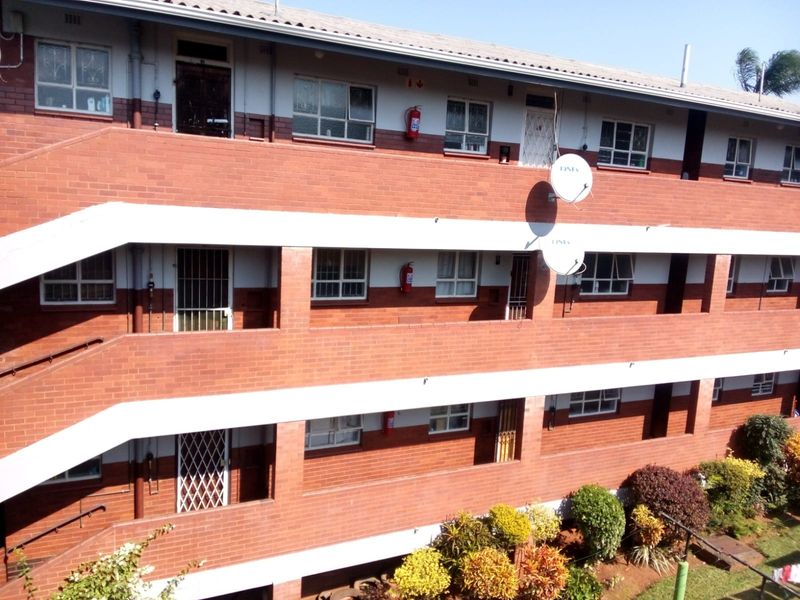 Exterior view of a multi-story apartment building featuring red brick facade, external staircases, and security gates on unit entrances.