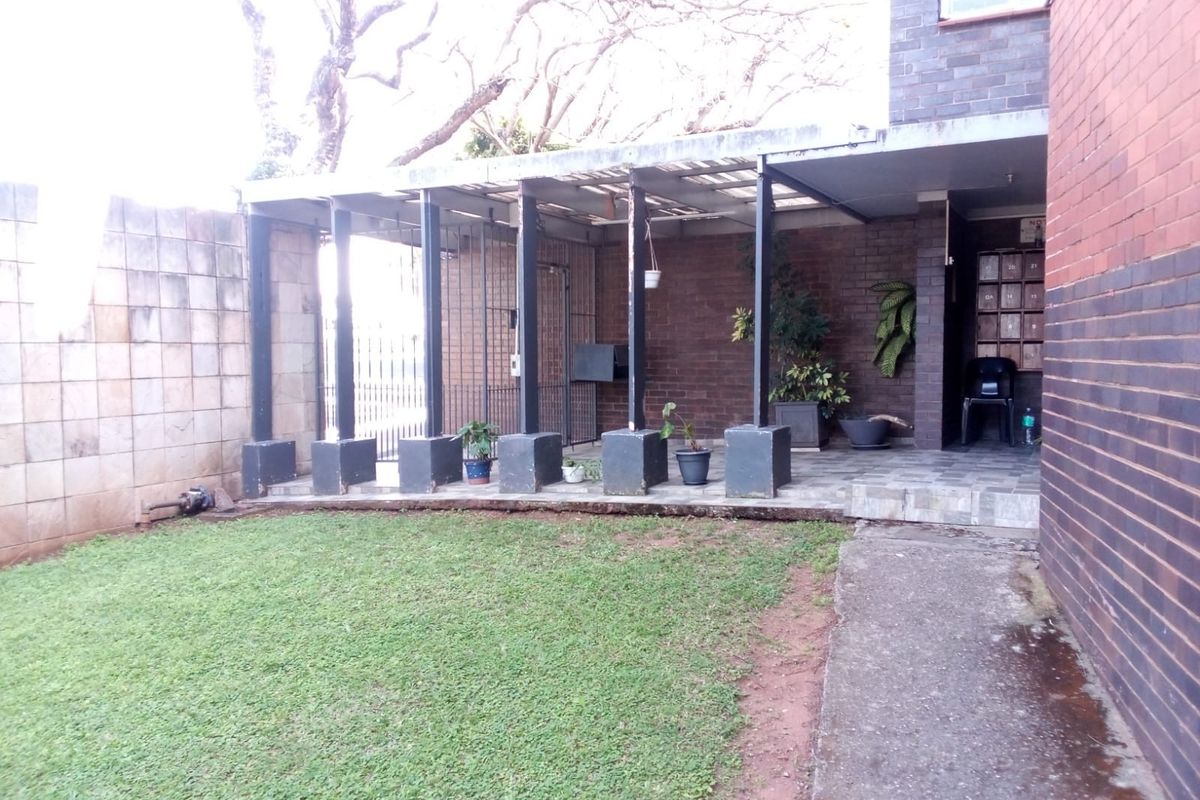 A rear exterior view of the apartment featuring a covered patio area, brick facade, paved walkway, and a small private lawn.