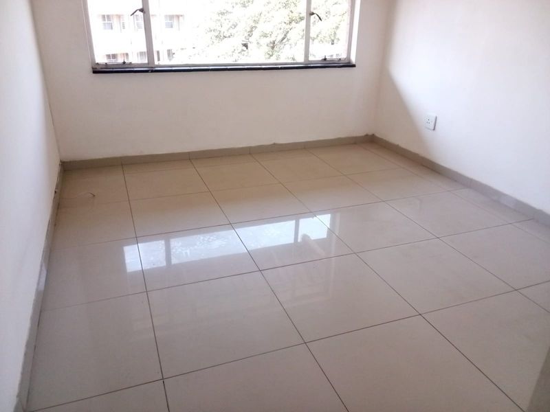 A bright, empty bedroom featuring neutral tiled flooring, clean white walls, and a large window providing natural light.