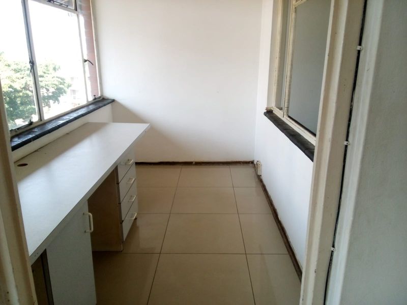 A sunlit enclosed balcony or study area featuring tiled flooring, a built-in desk, and large windows with stone sills.