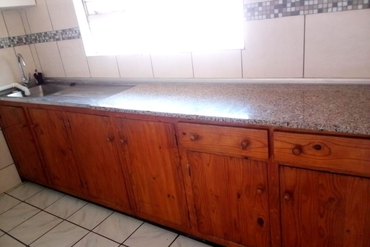 A functional kitchen featuring wooden cabinetry, granite countertops, a stainless steel sink, and tiled backsplash with neutral floor tiles.