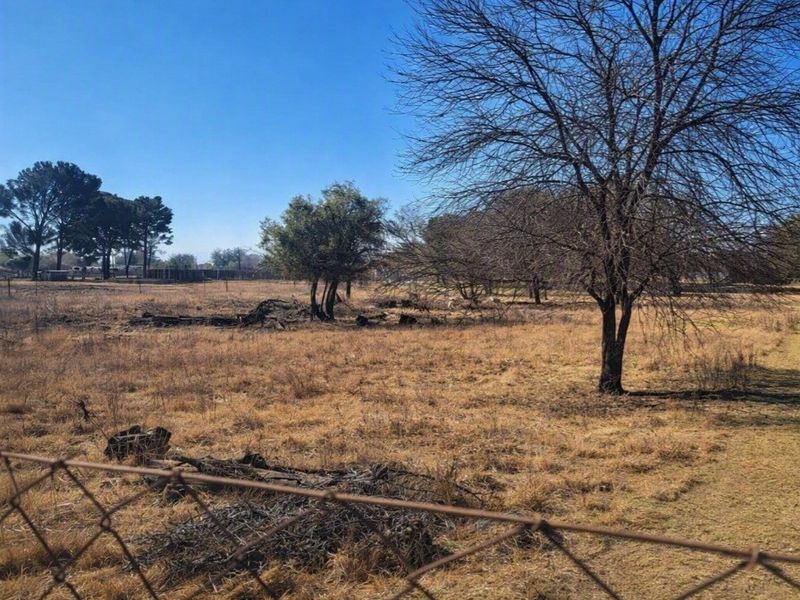A view of a vacant residential plot featuring dry grass, mature trees, and perimeter wire mesh fencing.