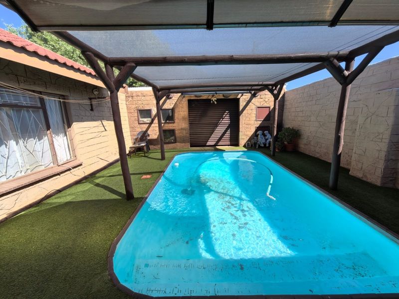 Outdoor pool area featuring a swimming pool covered by a timber pergola and shade sail, surrounded by artificial turf and high boundary walls.