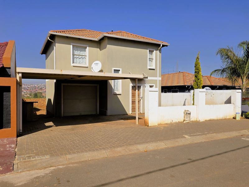 Front view of a two-story residential house featuring a paved driveway, attached carport, roller shutter garage door, and perimeter boundary wall.