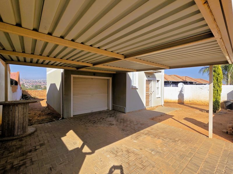 A paved driveway area featuring a covered carport structure and a single roller shutter garage door, set within a fenced residential property.