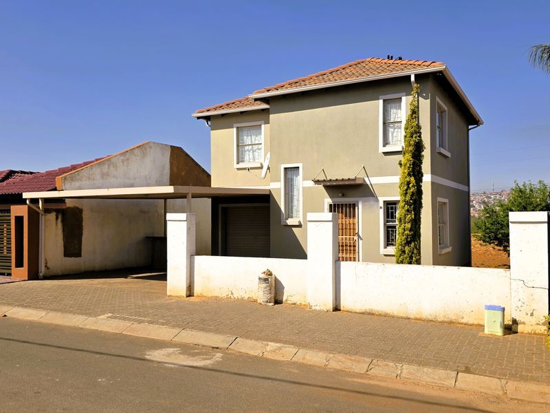 Front view of a two-story residential house featuring a paved driveway, attached carport, and perimeter boundary wall.