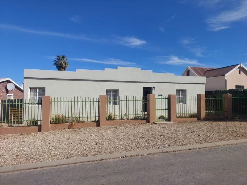 Front elevation of a single-story house featuring a parapet roofline, secured by a perimeter fence with brick pillars and a metal gate.